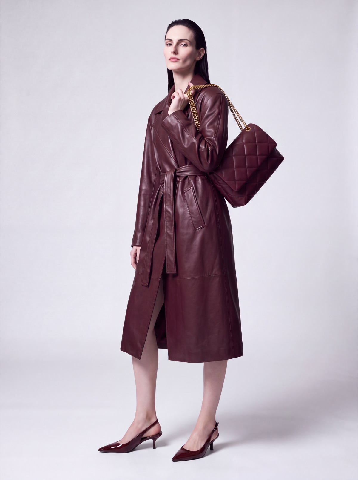 Burgundy leather trench coat on the same AI model, three-quarter view with the quilted bag on the shoulder