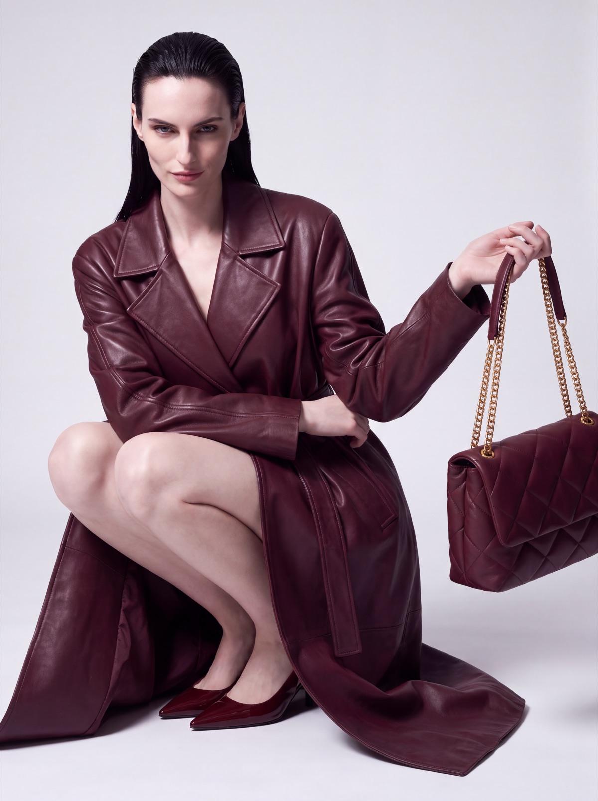 Burgundy leather trench coat on the same AI model, crouching with the quilted bag