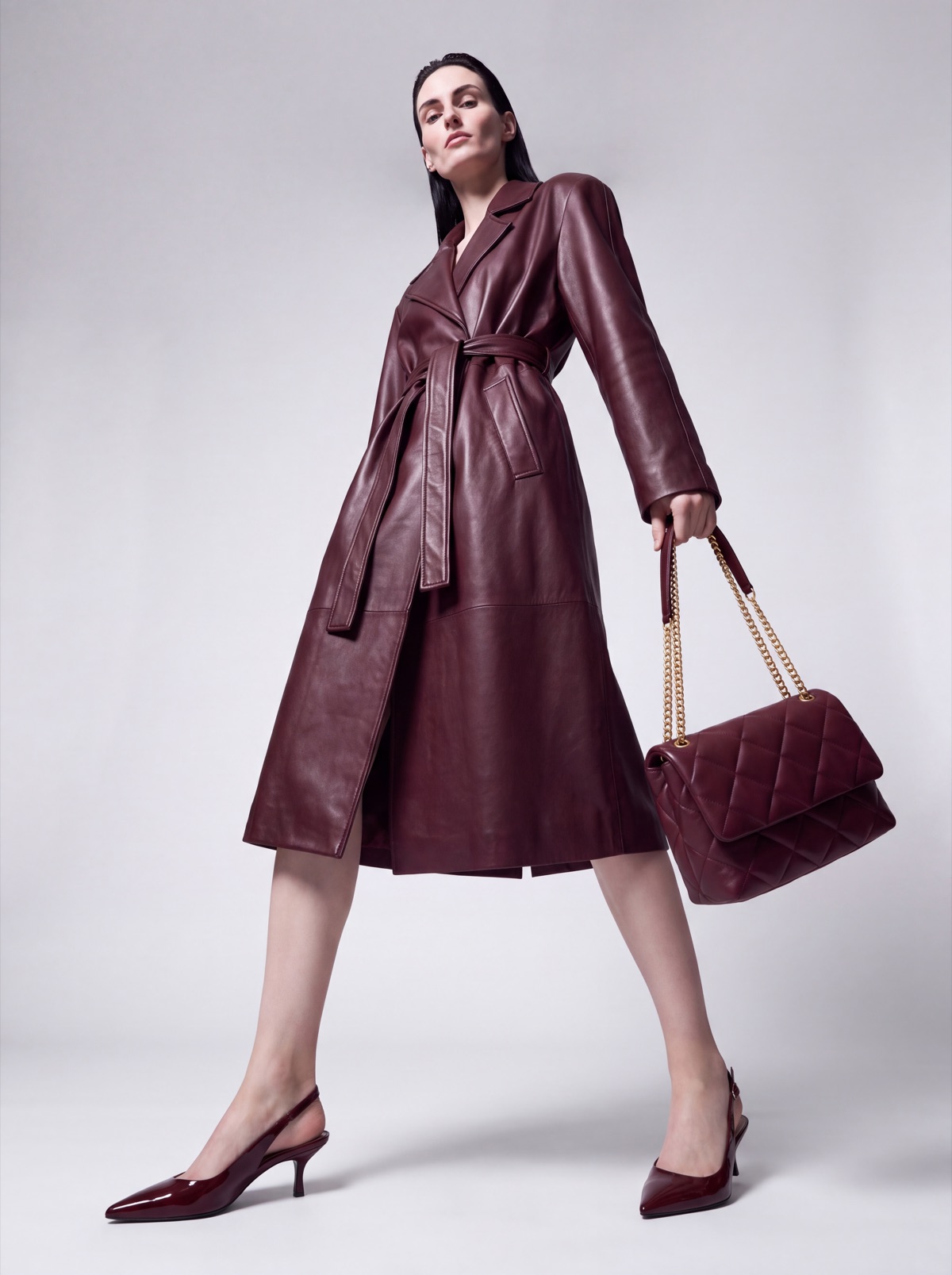 Burgundy leather trench coat and quilted bag on an AI model, walking three-quarter studio angle